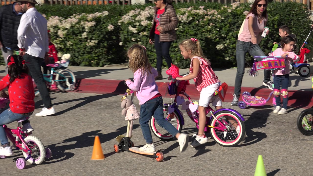 Children ride small bicycles and scooters near orange cones on a sunlit street in Cabo San Lucas, with adults walking nearby and hedges behind them.