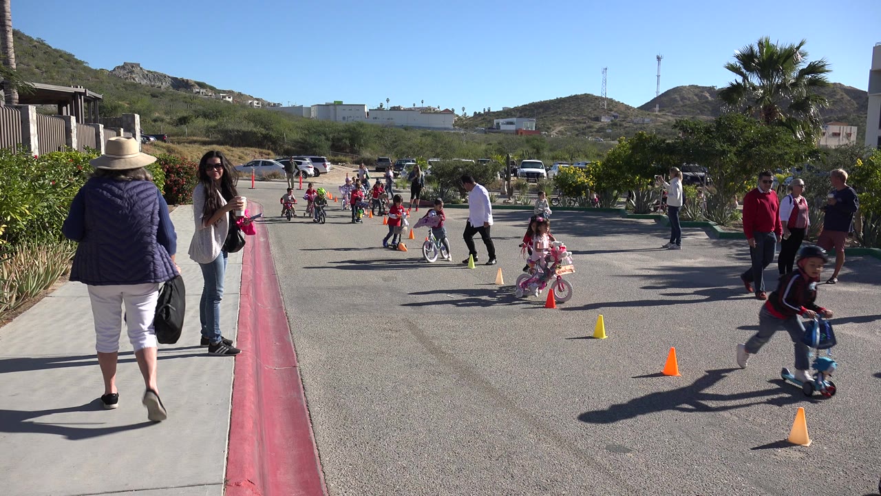 Children riding bicycles around cones while adults watch beside a sunny road.