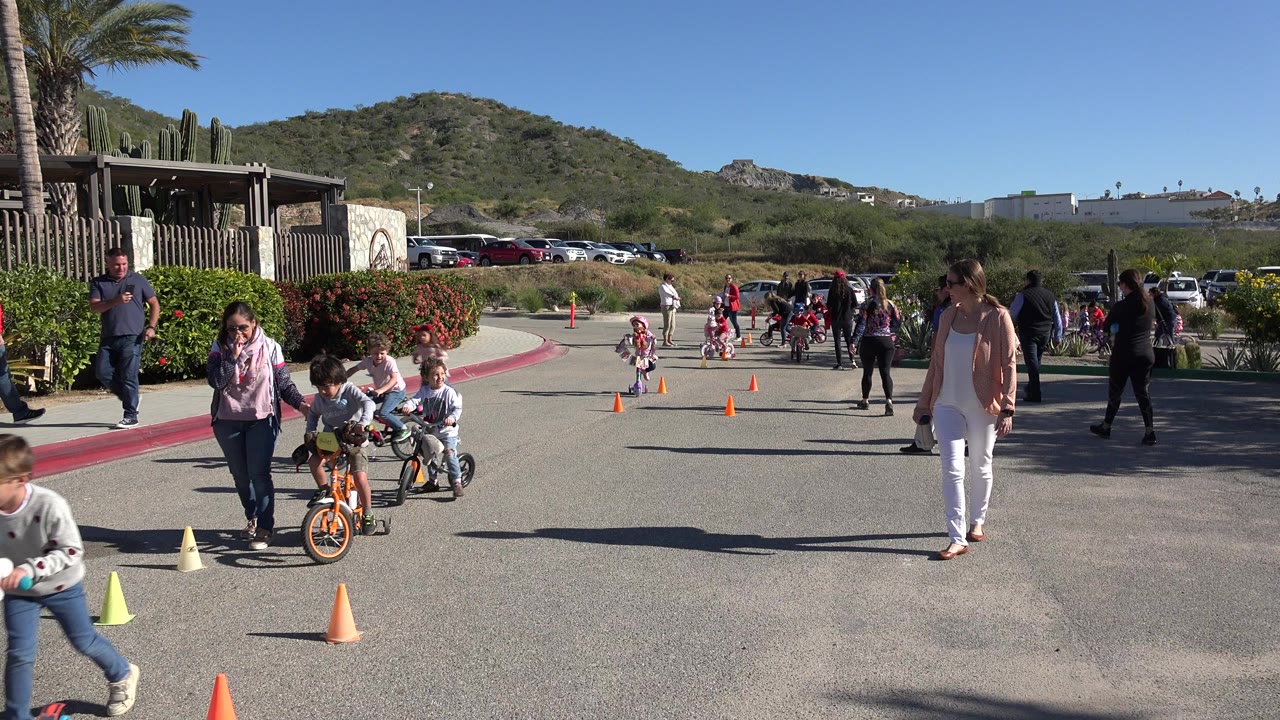 Children ride small bikes through cones while adults watch on a sunny paved lot.