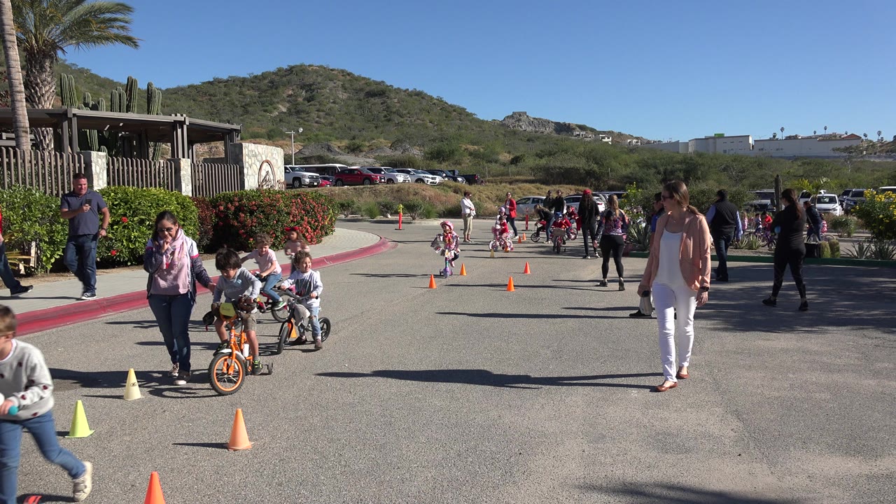 A child rides an orange bicycle near the foreground cones in Cabo San Lucas while more pupils and adults gather along the paved course.