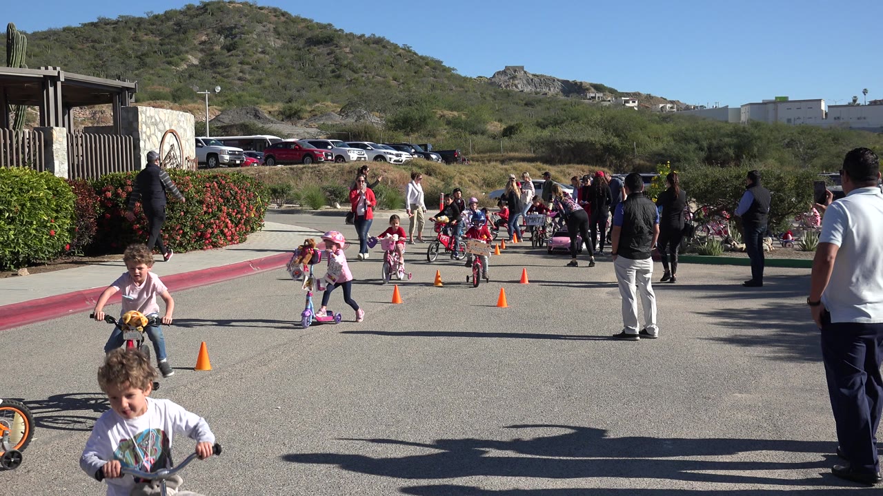 At the start of the Cabo San Lucas clip, children on small bikes and scooters approach orange cones on a paved course as adults stand along the sides.
