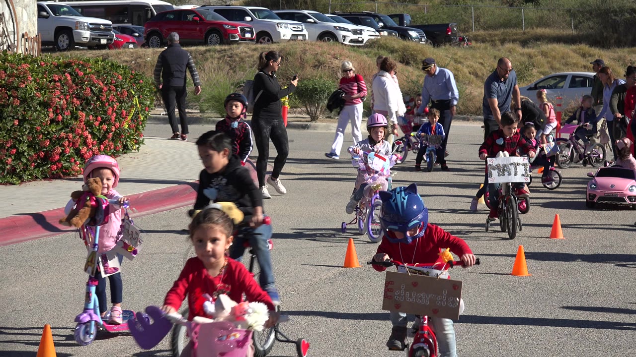 Children ride small bikes and scooters on a cone-marked outdoor course in Cabo San Lucas, Mexico, with adults supervising and parked cars behind them.