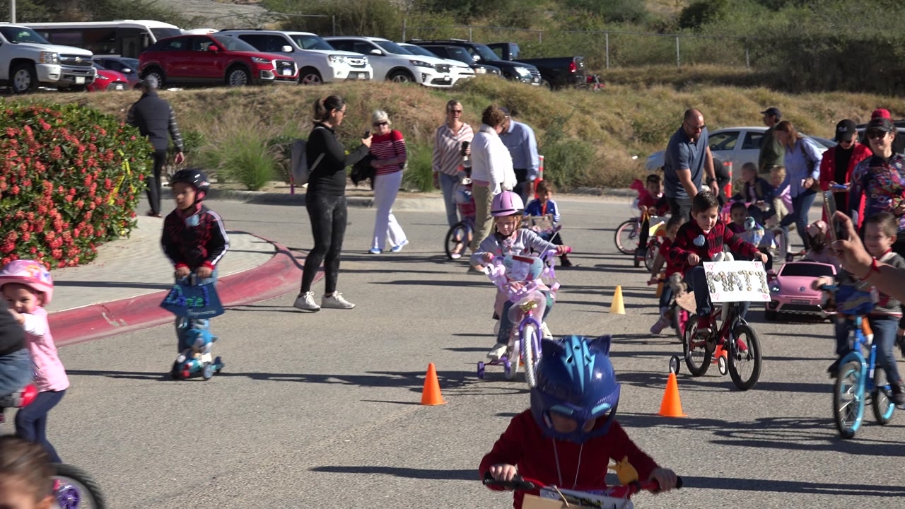 Children ride across a paved cone course with adults and parked vehicles in the background.