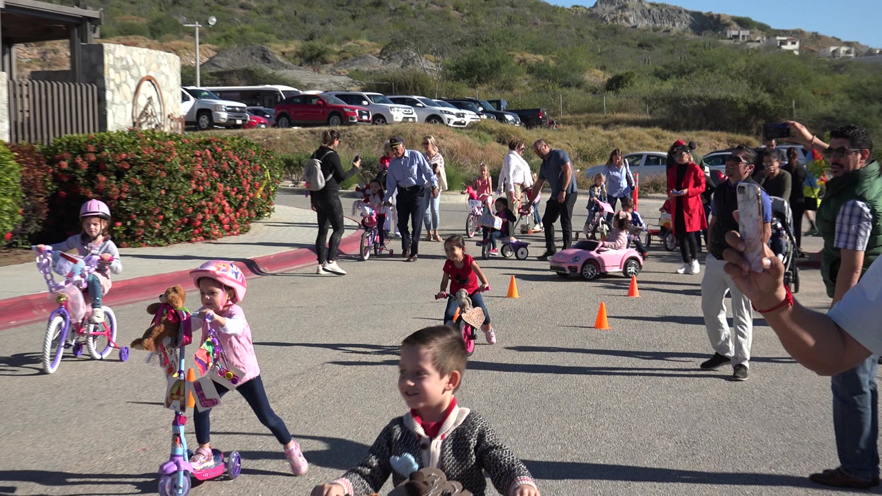 Children on scooters and bikes pass cones while adults watch near shrubs and hills.