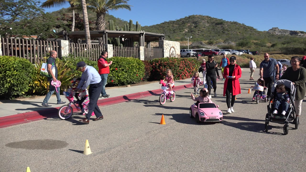 Children ride bikes and a toy car on a sunny street with adults nearby.