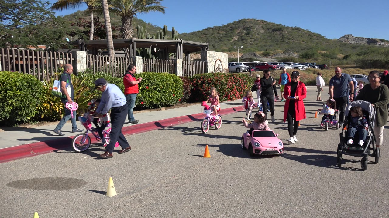 A Cabo San Lucas street scene shows an adult bending near a child's bicycle, children moving on scooters and a toy car, and parents standing along the red curb under bright sun.