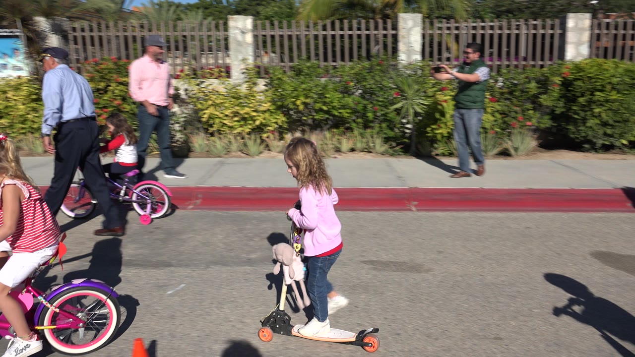 In Cabo San Lucas, Mexico, children move past the camera on a pink bicycle and a scooter while adults stand near a red curb and landscaped fence.