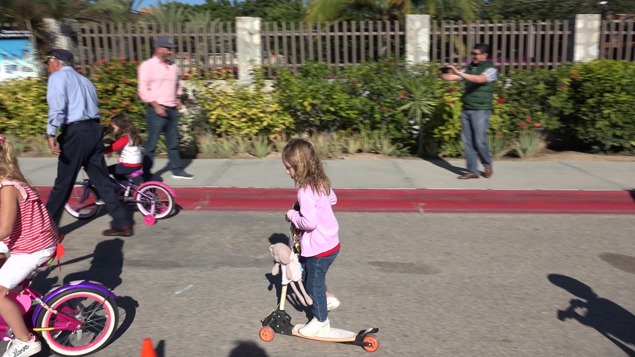 In Cabo San Lucas, Mexico, a girl on a pink bicycle and another on a scooter pass in front of adults standing along the curb under strong sun.