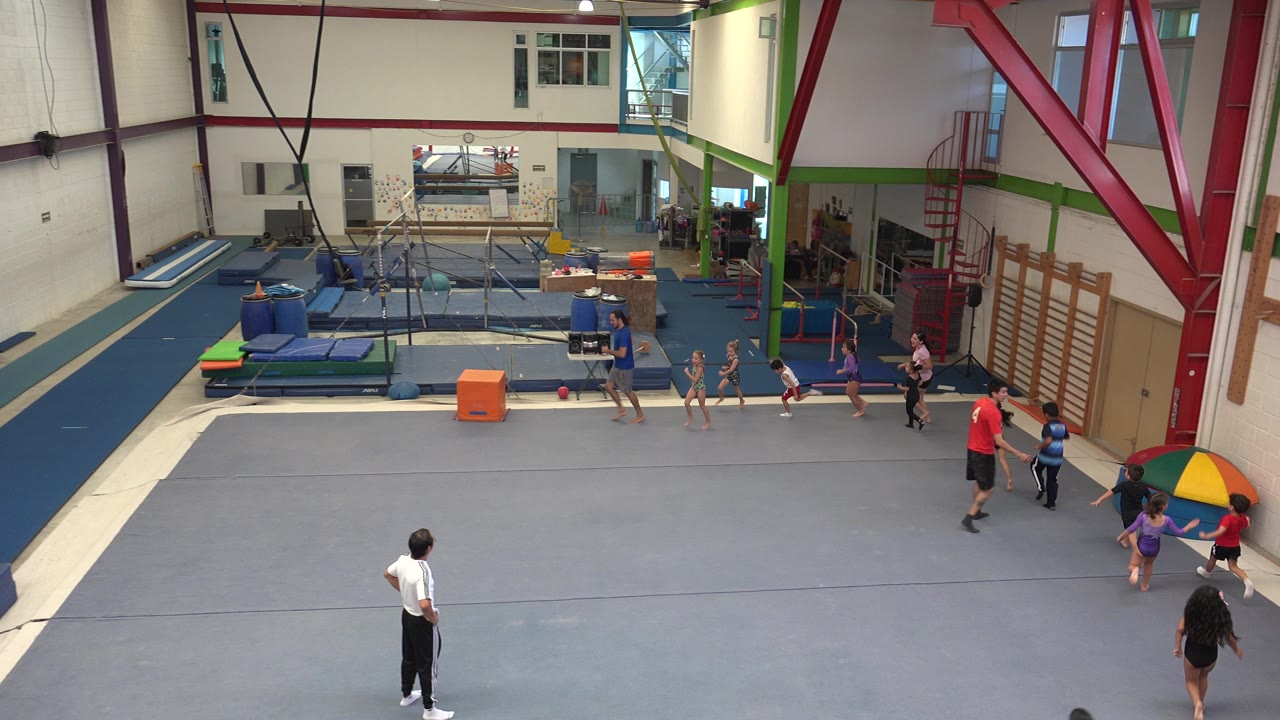 Wide high-angle view in Cabo San Lucas, Mexico, showing children lined up on a gray gym floor with adult instructors, blue mats, bars, and colorful pads in the background.