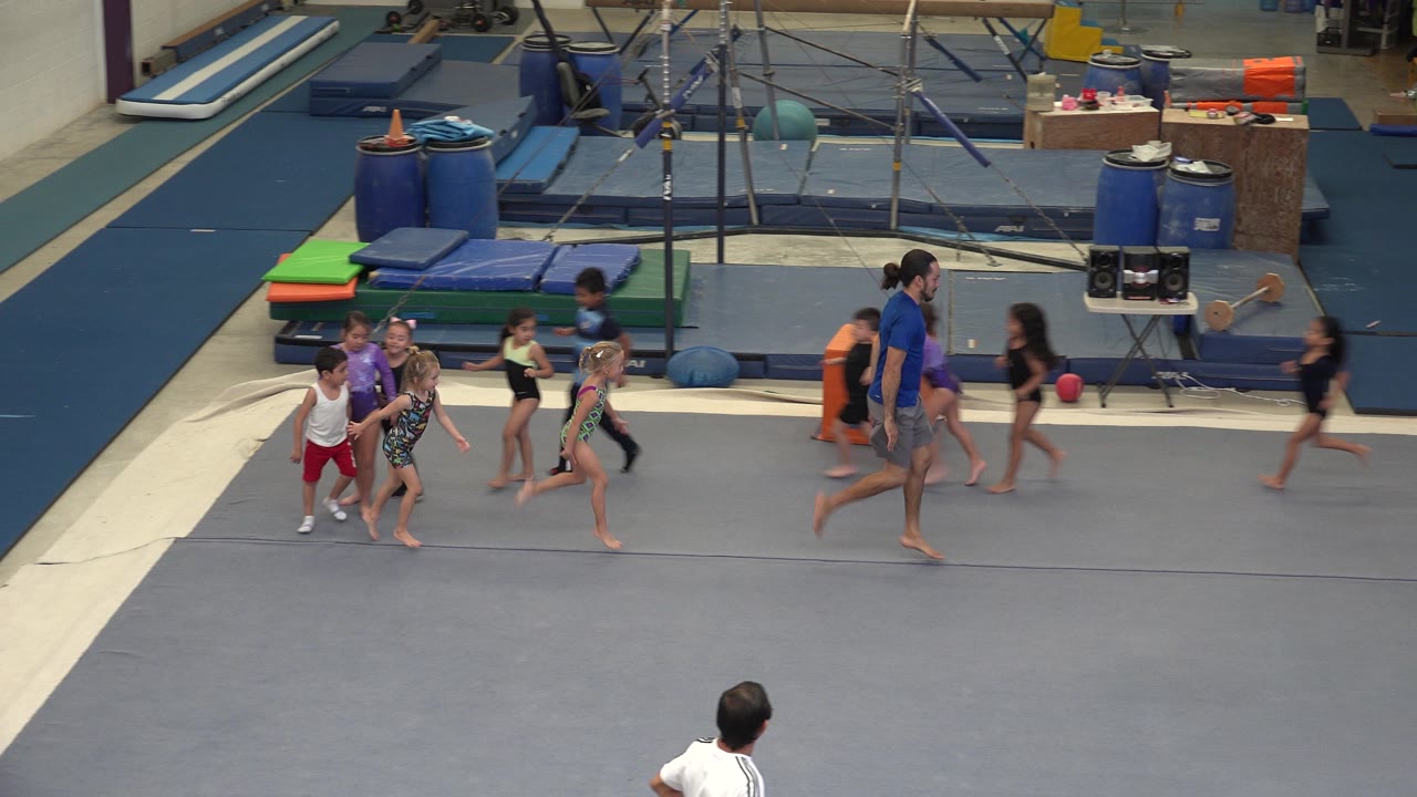 In Cabo San Lucas, children in athletic clothes move across a gray gymnastics floor behind a coach, with mats and training equipment in the background.