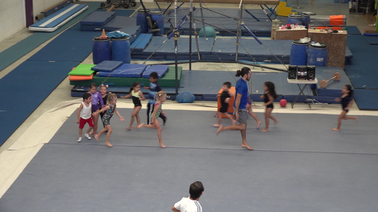 In Cabo San Lucas, young gymnasts continue behind an adult coach while another child runs ahead on the same gray floor, with stacked mats and equipment behind them.