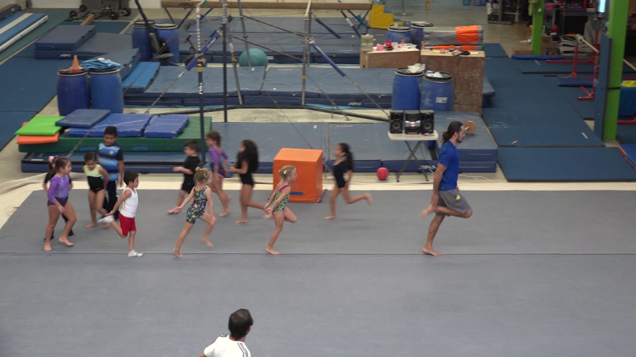 A coach leads children past gym mats and equipment.