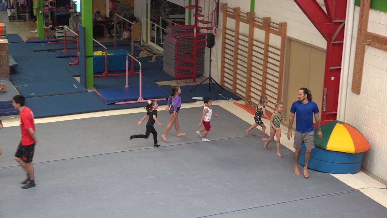 In Cabo San Lucas, Mexico, the running line of children follows the instructor past wall-mounted bars and mats, with the group spread across the center of the gym floor.