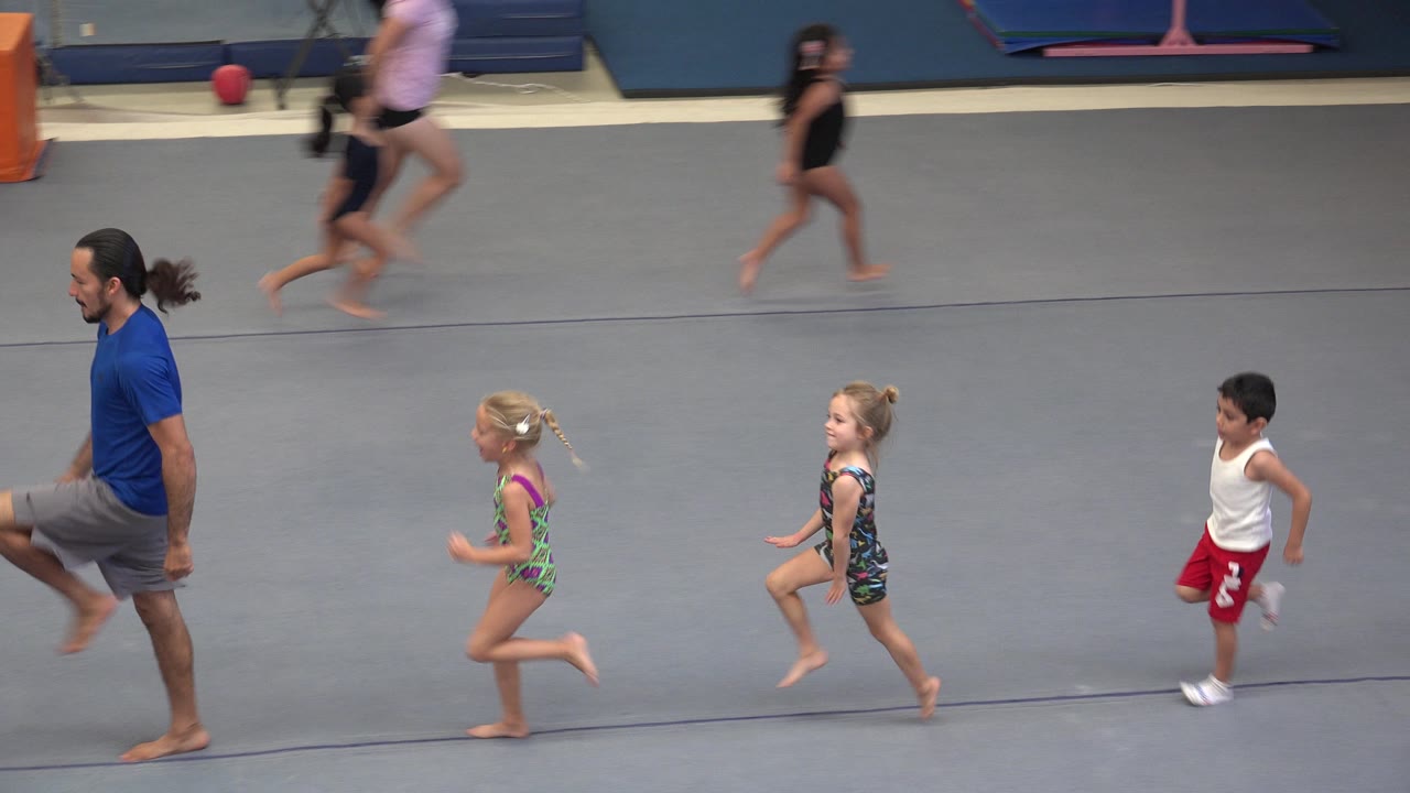 At 0:00 in Cabo San Lucas, Mexico, an adult instructor in a blue shirt leads young children running across a gray gymnastics mat while other children move in the background.