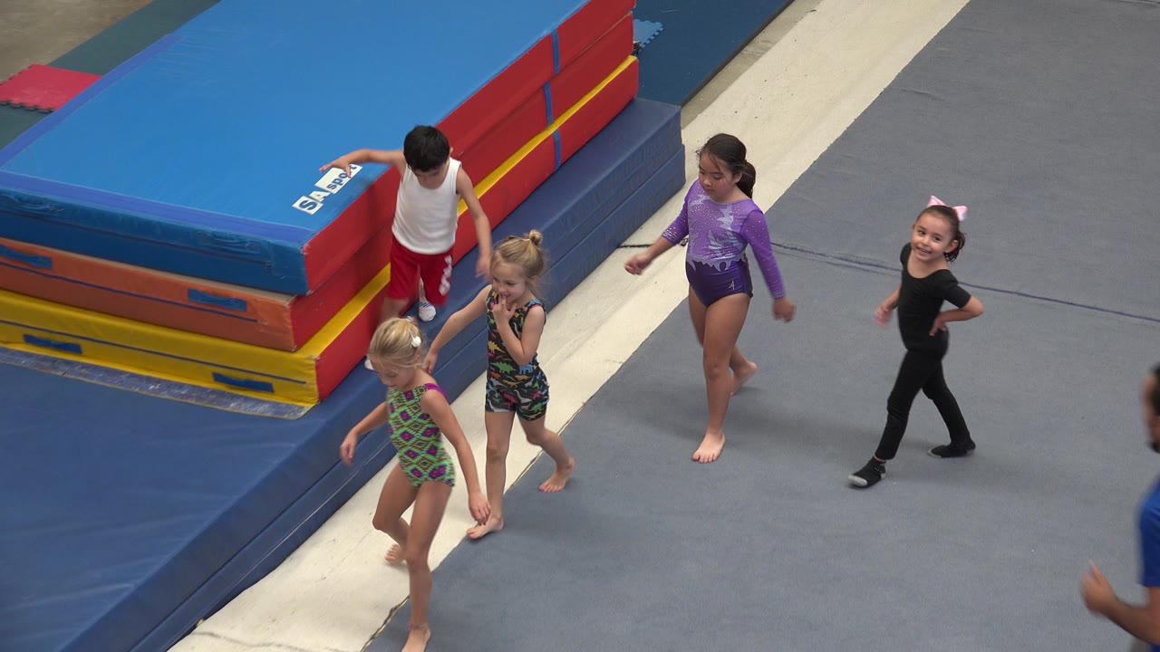 Children in athletic outfits walk across a gray padded gym floor in Cabo San Lucas, Mexico, with stacked red, yellow, orange, and blue mats at the left edge.