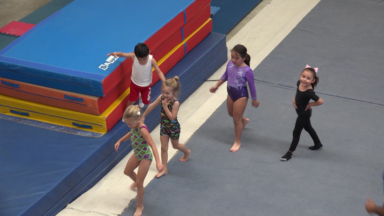 A small group of children continues across the padded gym floor in Cabo San Lucas, Mexico, with colorful stacked mats filling the upper left of the frame.