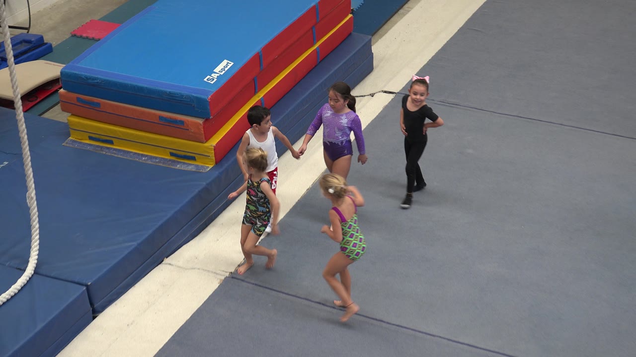 Children move farther across the gym floor near a hanging rope and mats.