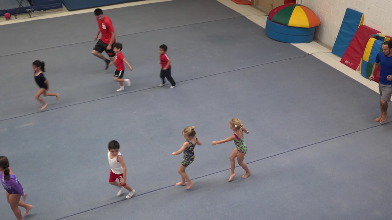 Young gymnasts continue across the padded floor as a coach in red and a coach in blue stand near the colorful gym mats in Cabo San Lucas, Mexico.