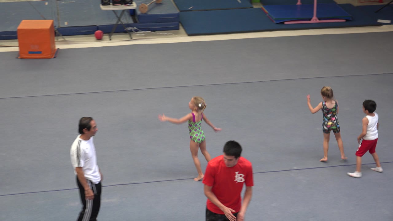 Children in athletic outfits warm up on a gray gym floor in Cabo San Lucas while two adult men stand near them and blue mats line the back wall.