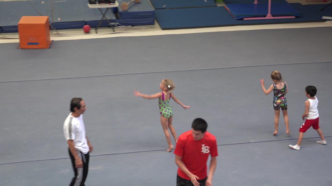 A blonde child in a green leotard gestures on the gray floor in Cabo San Lucas as another child and two adult men remain in view near the gym equipment.
