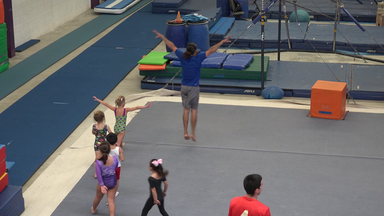 In Cabo San Lucas, children stand on a gray gym floor while an adult instructor in a blue shirt raises his arms near stacked mats and training equipment.