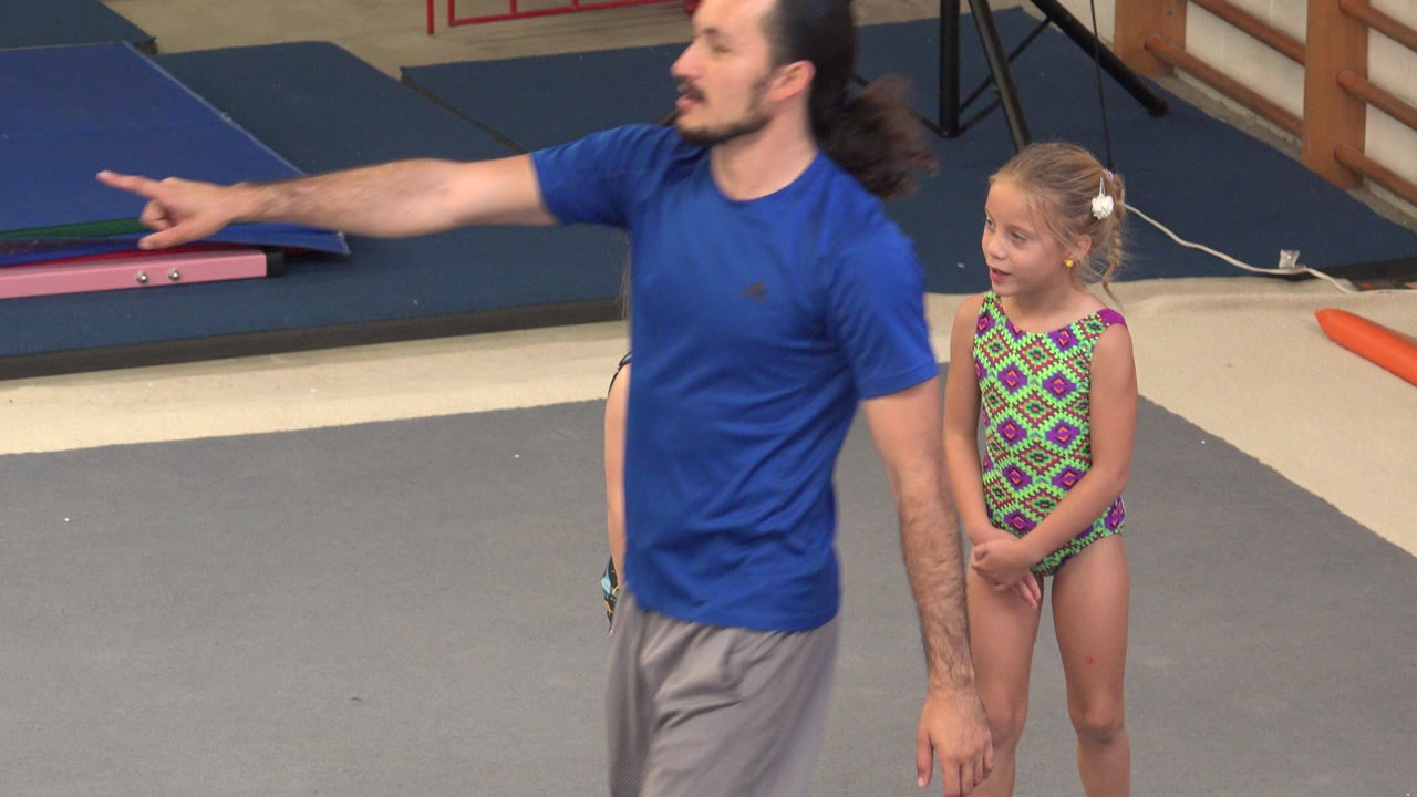 Adult instructor points while a young gymnast stands nearby.
