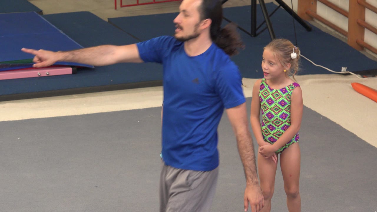 In Cabo San Lucas, Mexico, the instructor remains partly in front of a young gymnast in a patterned leotard, with blue mats and gym equipment behind them.
