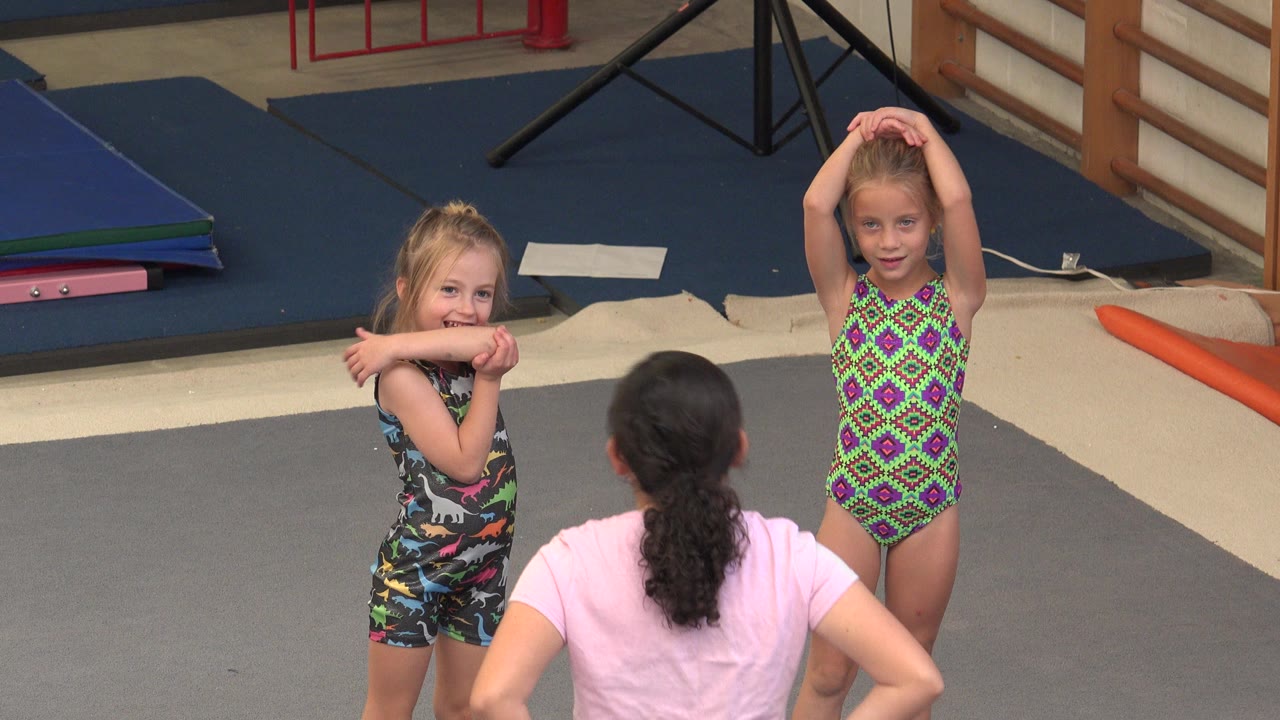 Two girls stretch on a gym mat while an instructor faces them.