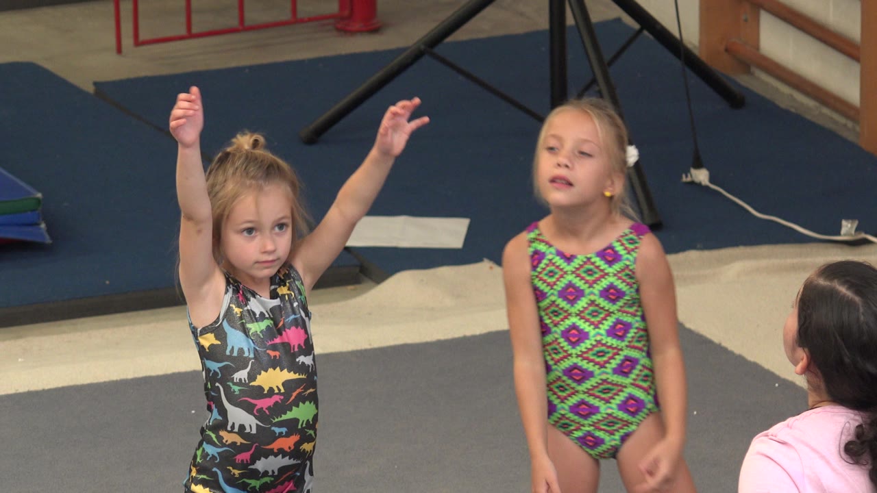 In Cabo San Lucas, one young girl keeps her arms lifted while another stands beside her in a patterned leotard and the adult coach remains partly seen from behind.