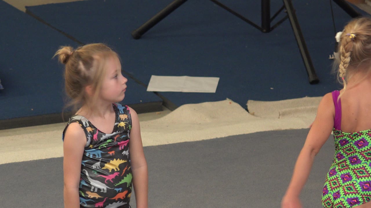 Young girl in a colorful leotard standing on a gym floor beside another child.