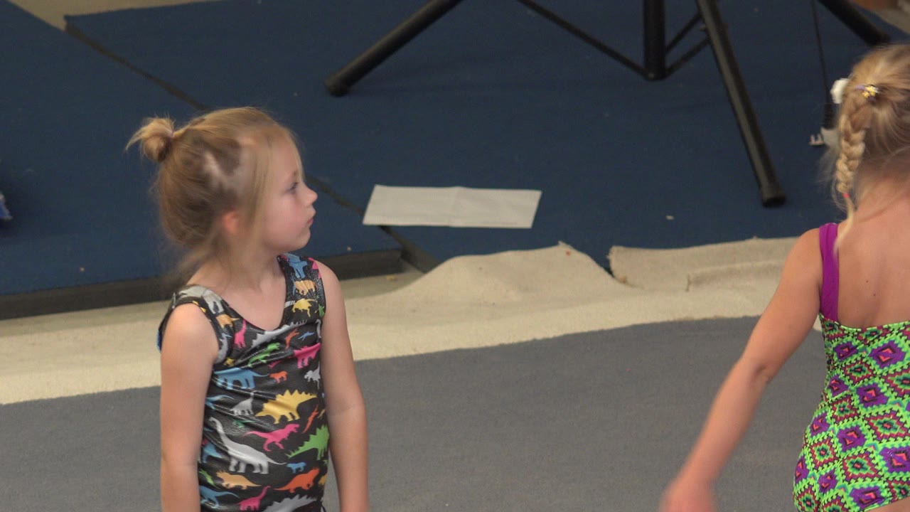 Young girl in a patterned leotard looks sideways in the Cabo San Lucas gym while another child's torso and arm are partly cropped at the right.