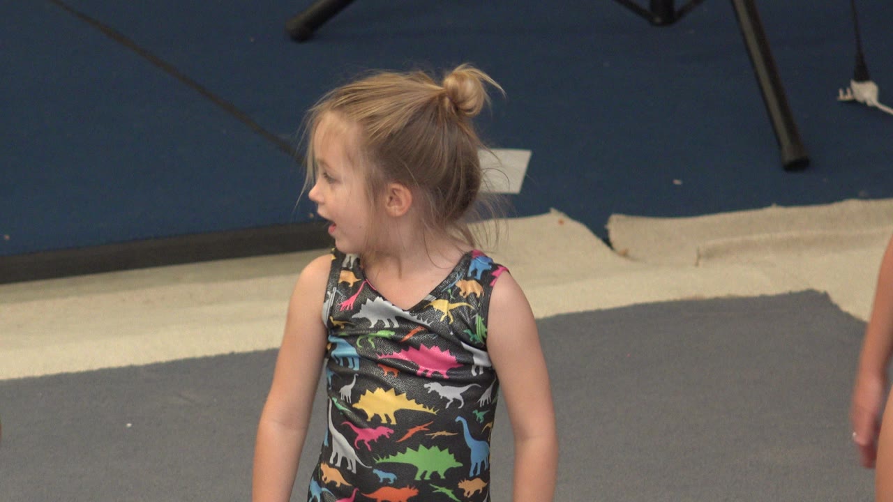 Blonde child in a colorful dinosaur leotard looking left on a padded floor.
