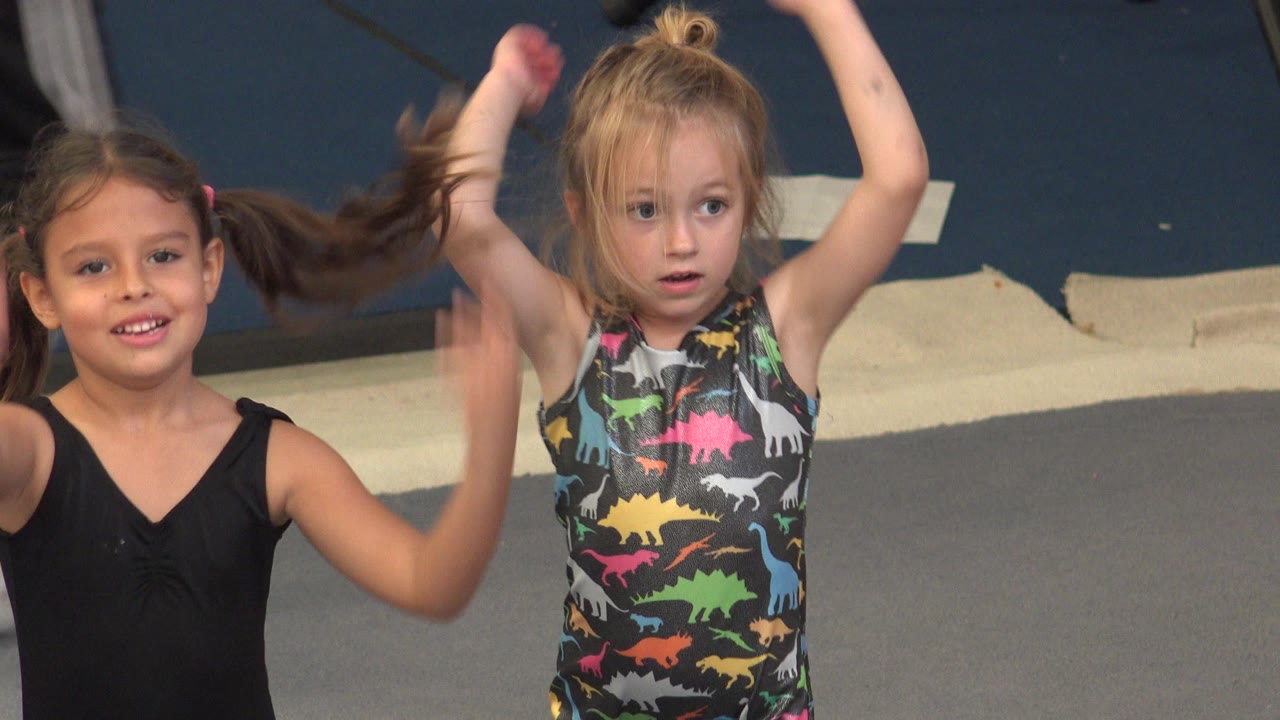 Two young girls in Cabo San Lucas stand on a gray padded floor with their arms lifted, one wearing black and the other in a colorful dinosaur-print leotard.