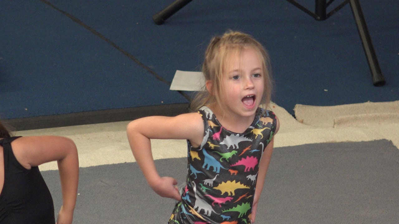 Blonde young girl in a colorful dinosaur-pattern leotard stands on gray gym mats in Cabo San Lucas, Mexico, with a partial child at the edge of frame.