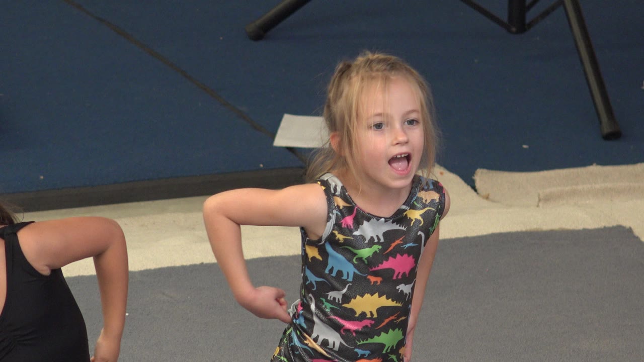 The blonde child faces forward with her mouth open during warmup, while another child is partly visible in the Cabo San Lucas gym setting.