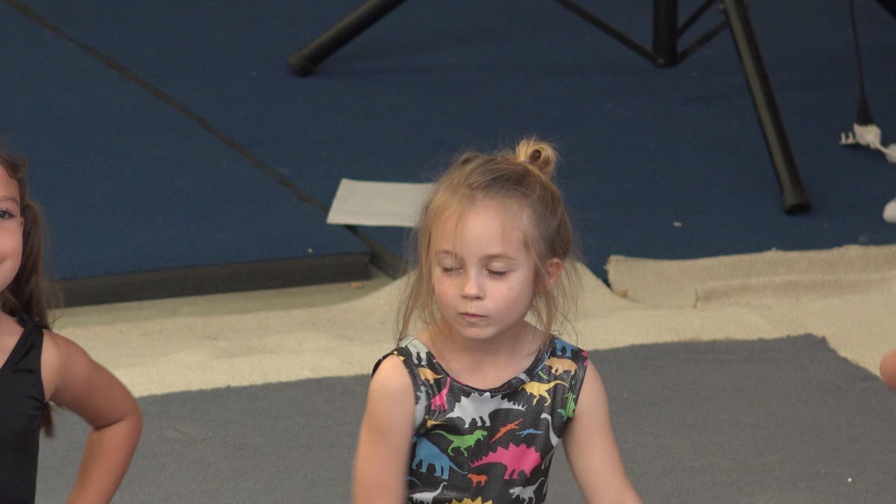 At 0:00 in Cabo San Lucas, Mexico, a young blonde girl in a dinosaur-print leotard stands on a gray padded floor while another child is partly visible at the left edge.