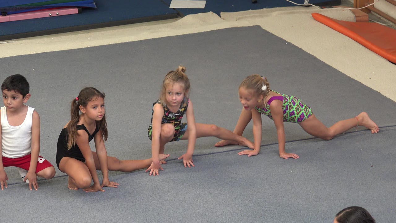 Young gymnasts stretch on a padded floor in an indoor gym.