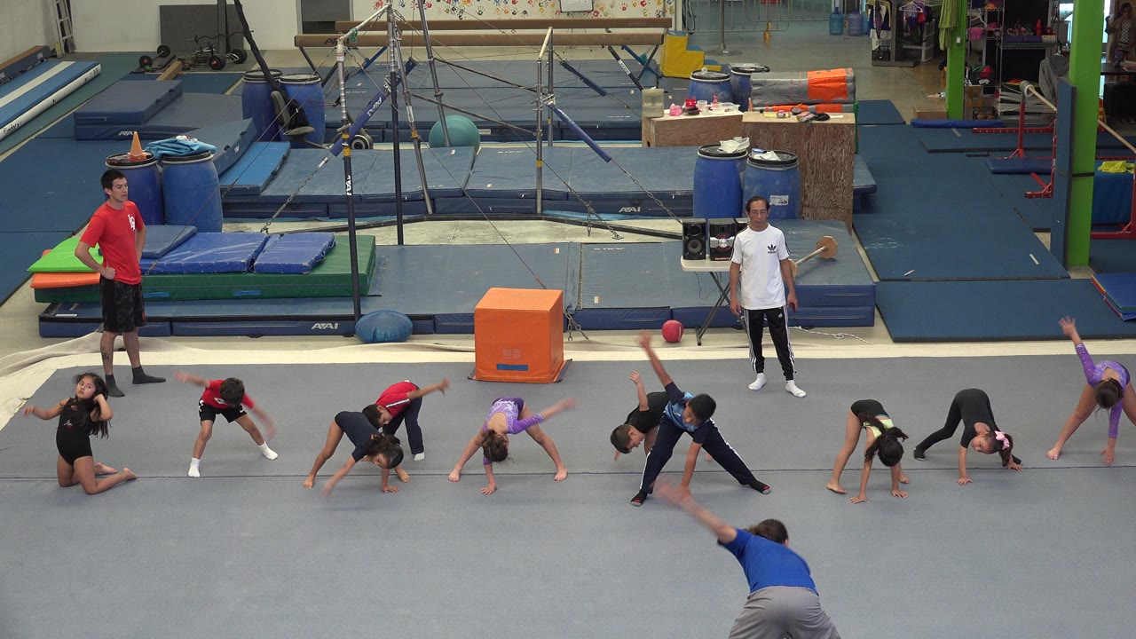 In Cabo San Lucas, Mexico, children stretch across a gray gym floor while two adult instructors stand near blue mats and gymnastics equipment.
