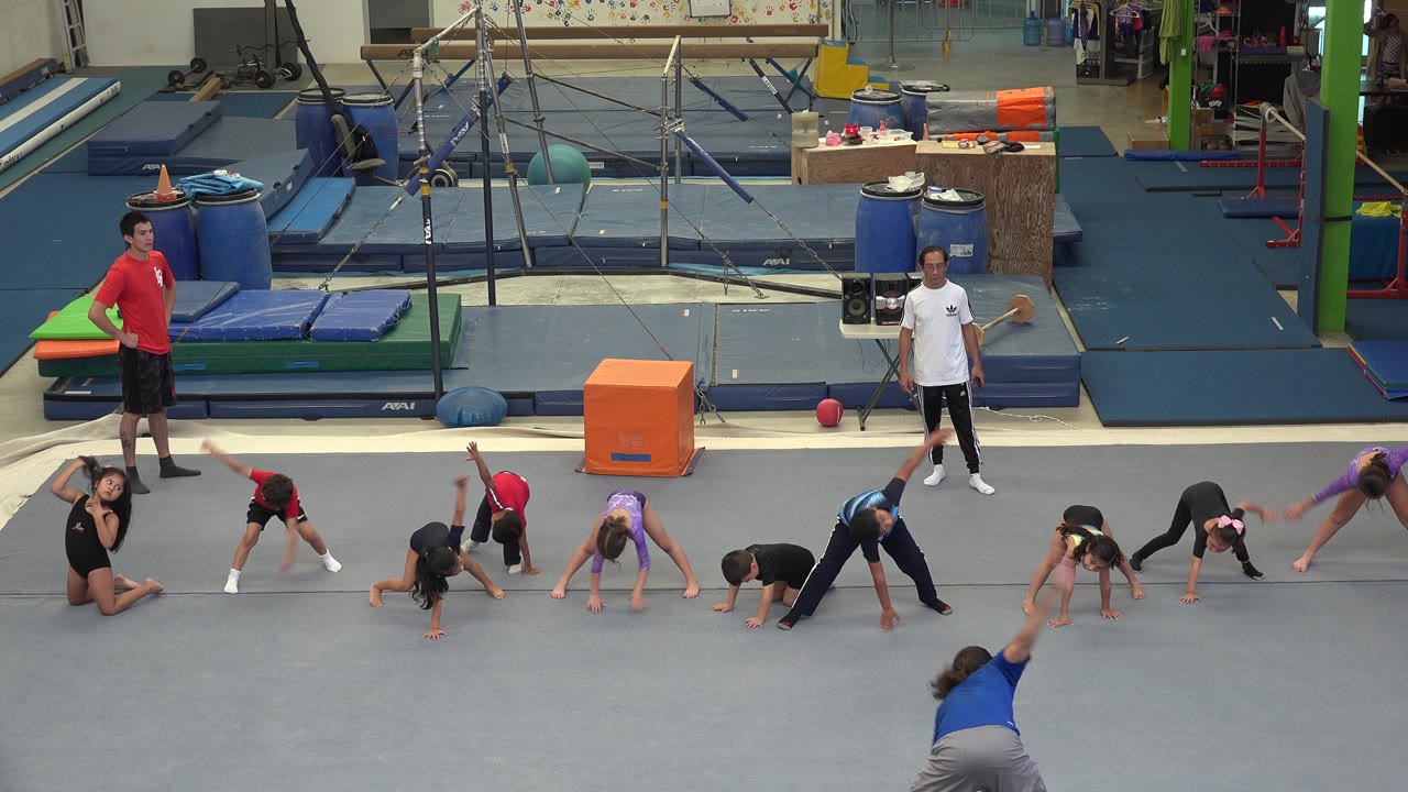 In Cabo San Lucas, Mexico, several children continue floor stretches while a child at the right edge raises an arm near the gym equipment area.