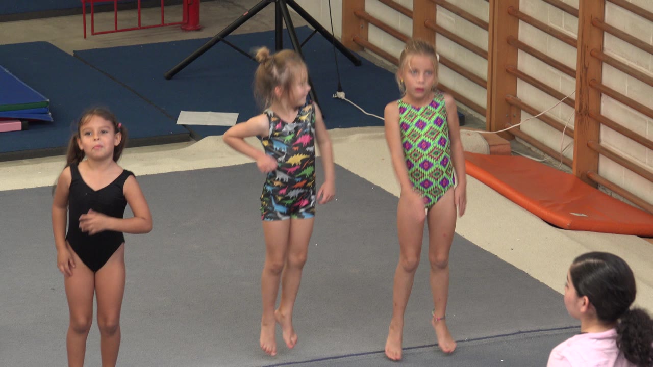 The children remain on the gym mat in Cabo San Lucas, with the girl in black facing forward and two girls in patterned leotards beside her.