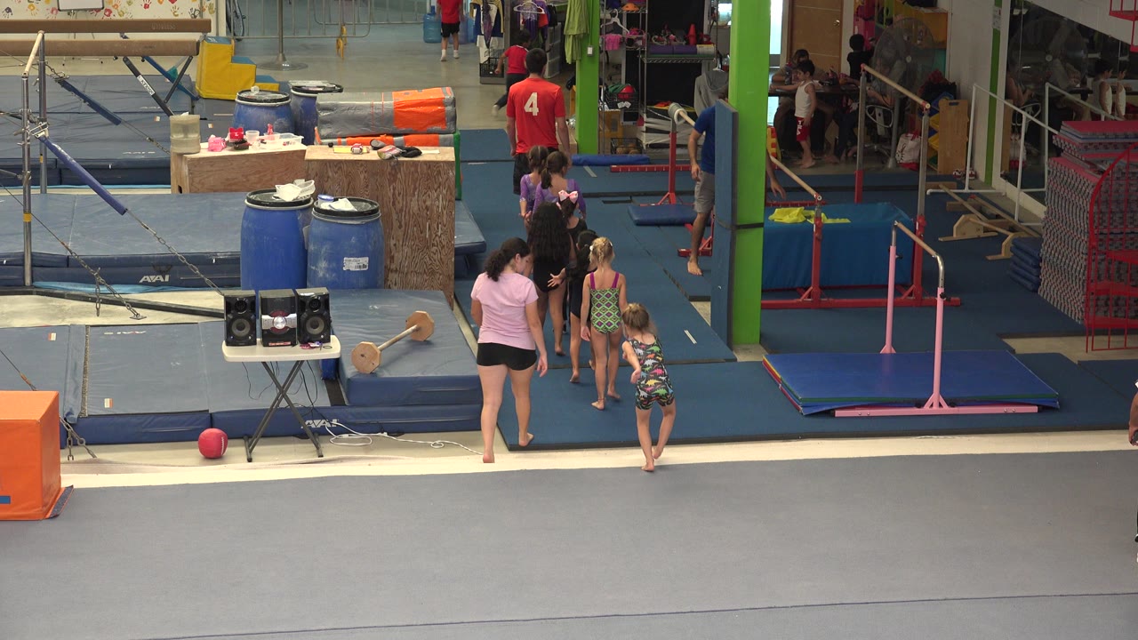 Wide view inside a Cabo San Lucas gymnastics facility, with blue mats, uneven bars, a green column, and adults and children gathered near the apparatus.