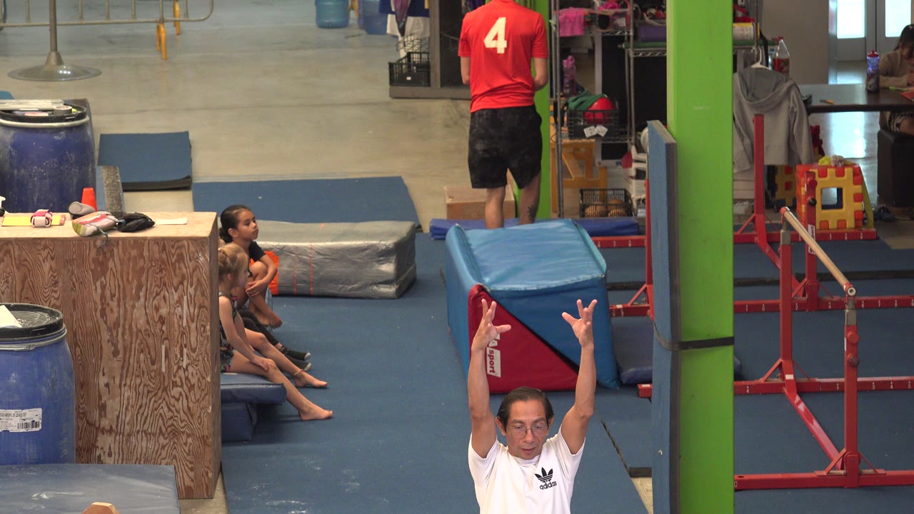 In Cabo San Lucas, Mexico, an indoor gym scene shows seated children on blue mats while an adult in a white shirt raises both arms at the lower edge of the frame.