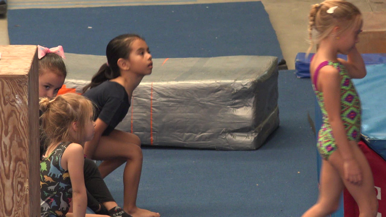 At 0:00 in Cabo San Lucas, Mexico, young girls in leotards sit and stand near blue flooring, a wood column, and gray padded mats in an indoor activity space.