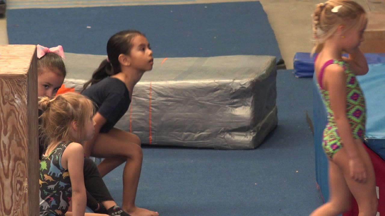 At 0:01 in Cabo San Lucas, Mexico, a girl in a colorful patterned leotard crosses the foreground while other children sit near a gray mat and wood column.