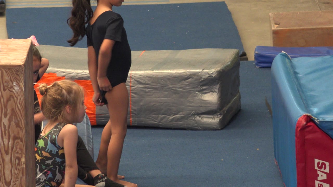 At 0:02 in Cabo San Lucas, Mexico, a girl in a black leotard stands near the mats while younger children remain seated beside the wood column and blue floor padding.