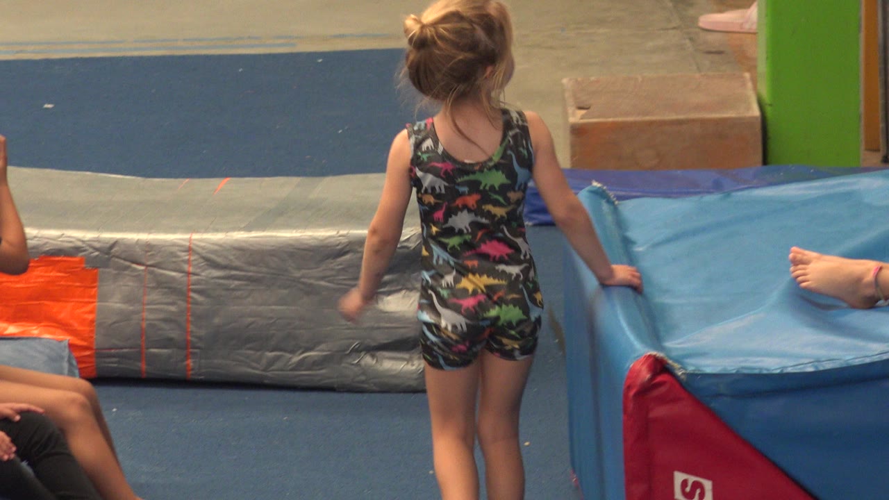 The child remains near the blue mat with one hand close to the padding, framed from behind in the indoor Cabo San Lucas space.