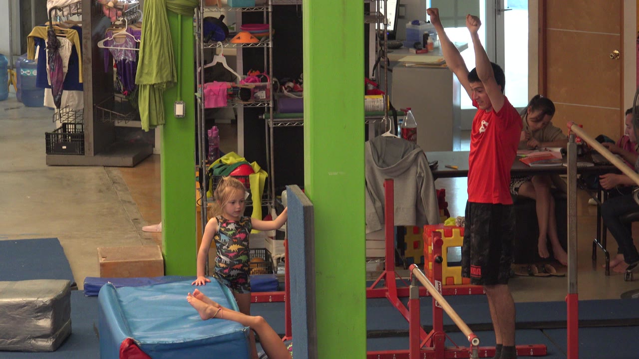 In Cabo San Lucas, Mexico, a young child sits on a blue gymnastics mat near low parallel bars while an instructor in a red shirt stands with arms raised beside a green column.