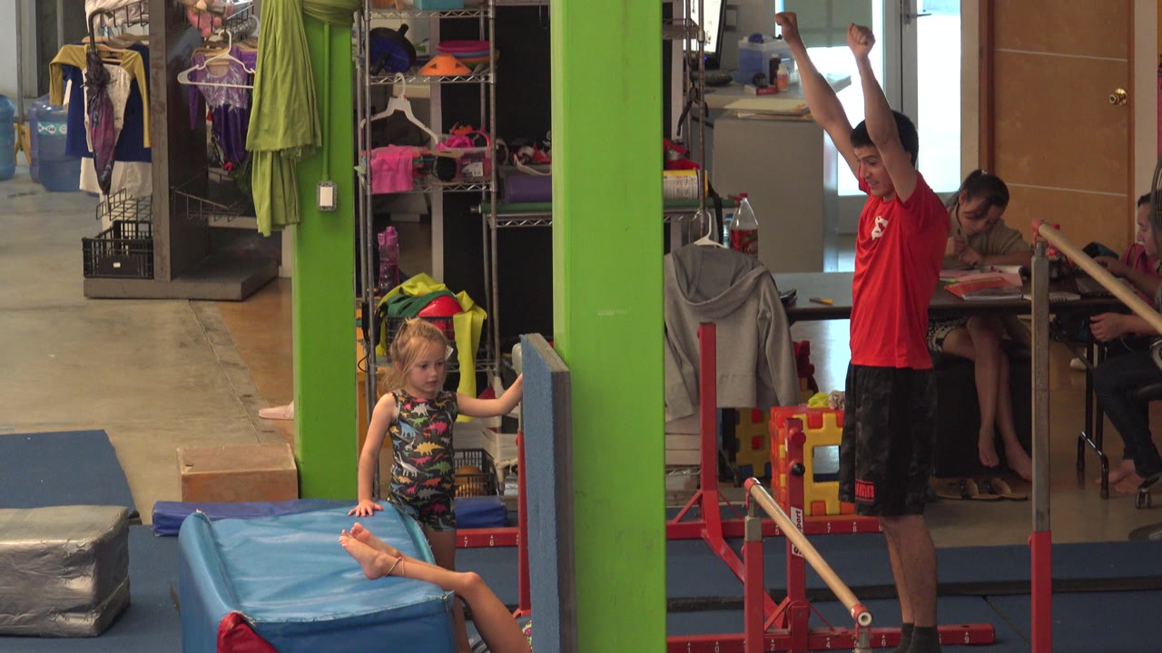 The instructor remains beside the parallel bars with both arms overhead while the child sits on the blue mat and adults are visible at tables in the background.
