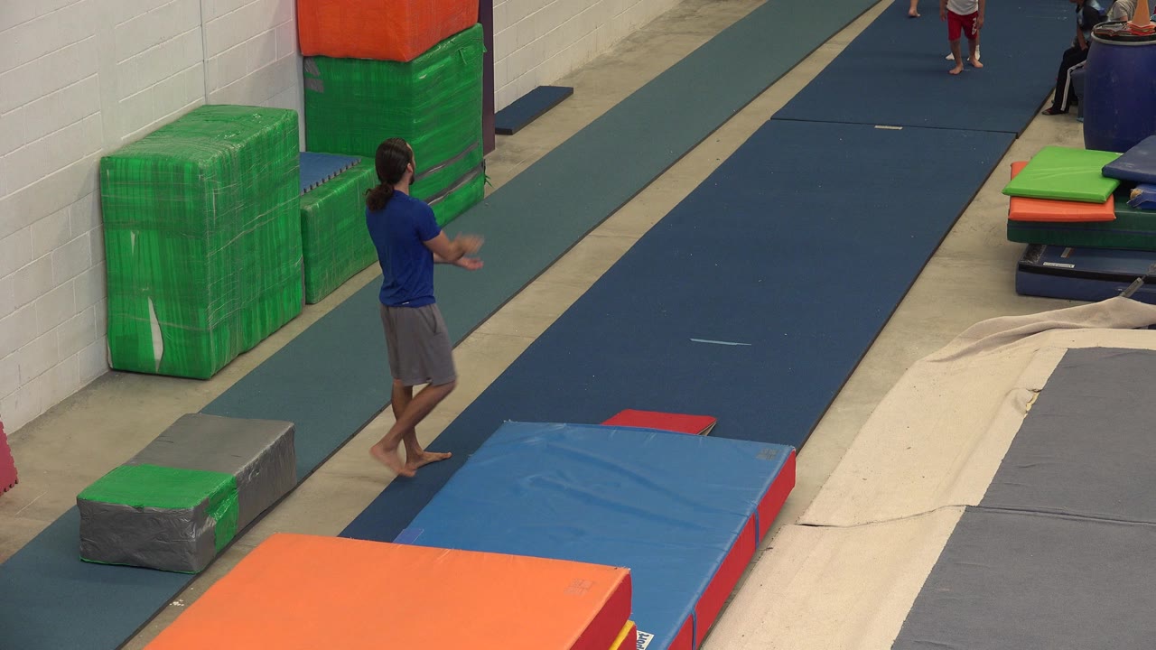 Adult man standing beside gym mats in an indoor training area.
