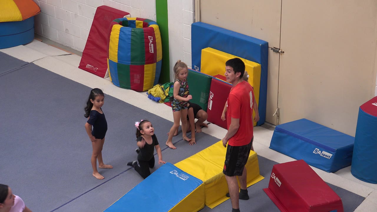 In Cabo San Lucas, Mexico, an instructor in a red shirt stands beside four children near blue and yellow foam mats and stacked padded equipment.